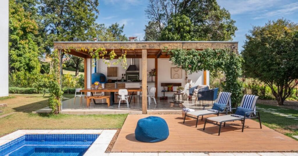 Charmosa área gourmet de casa tem pergolado florido, forno de pizza azul e piso em cerâmica